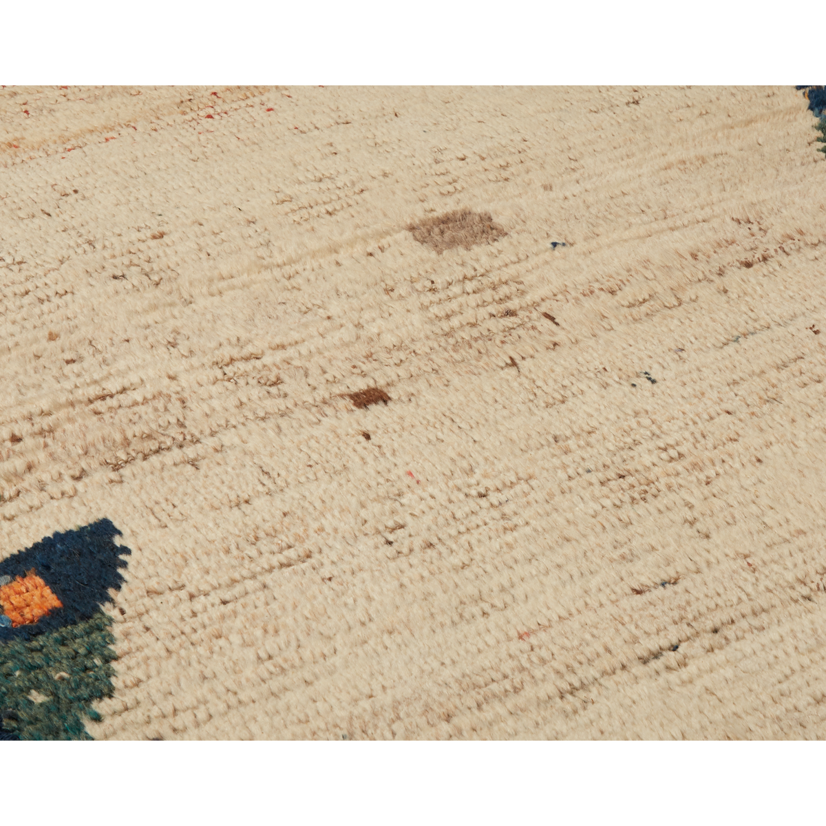 A close-up of a textured cream or off-white woven rug with subtle tonal variation and a small dark multicolor section visible in the lower-left corner.