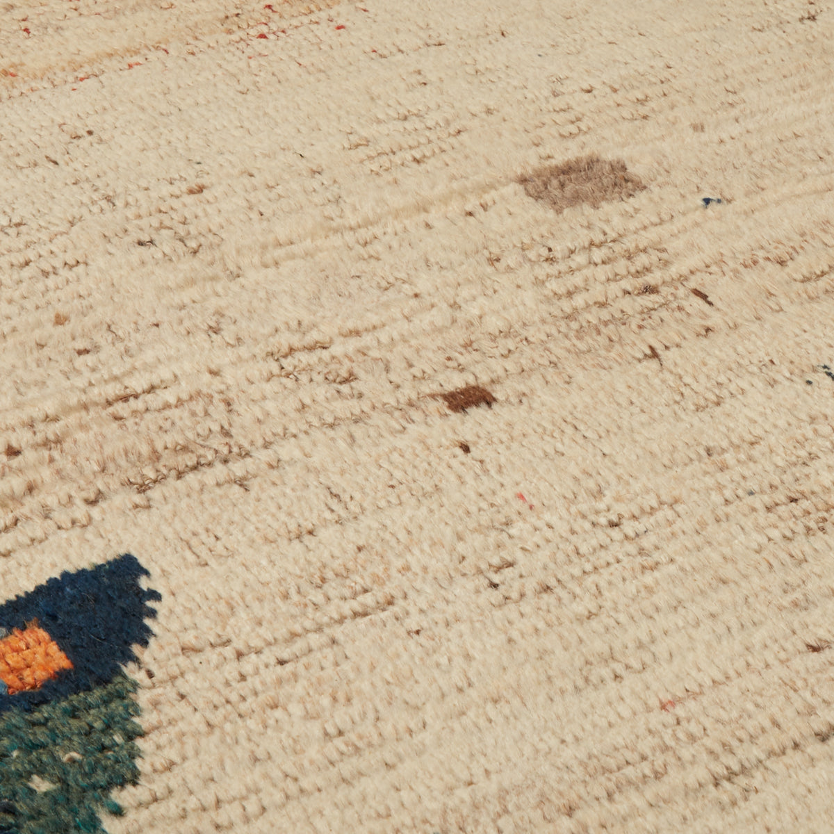 A close-up of a textured cream or off-white woven rug with subtle tonal variation and a small dark multicolor section visible in the lower-left corner.