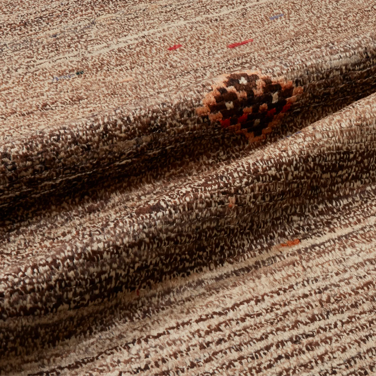 A close-up of a textured woven rug or textile in mixed brown, beige, and cream tones, with a small multicolor geometric accent visible near the center.