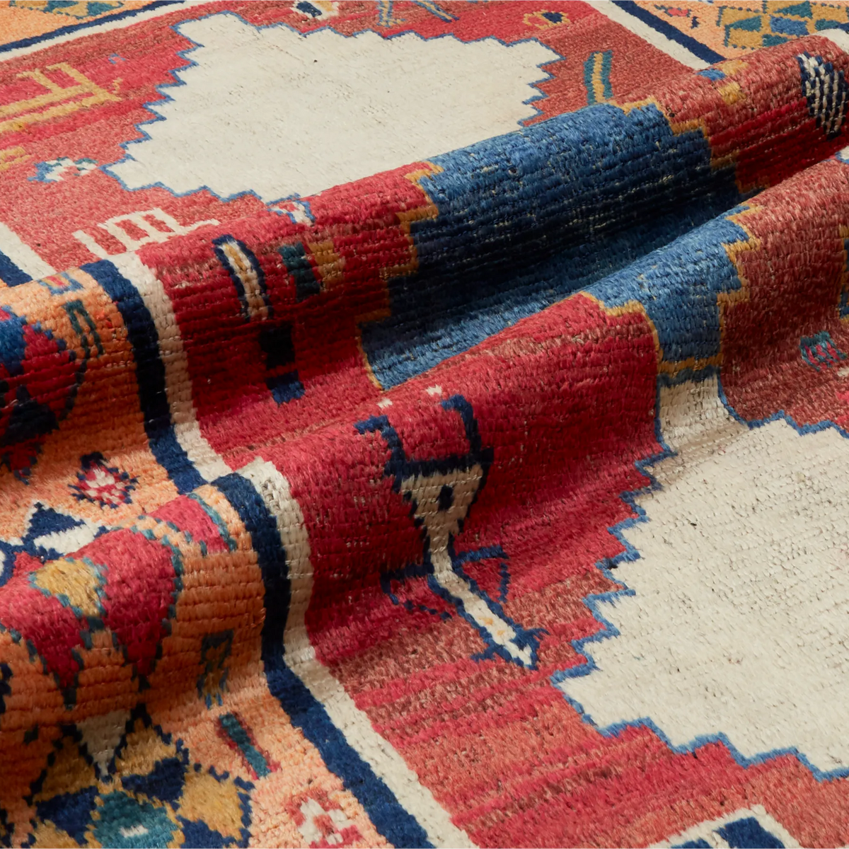 A close-up of a woven area rug with a traditional geometric pattern in red, blue, cream, and orange tones, shown with a folded section revealing the texture.