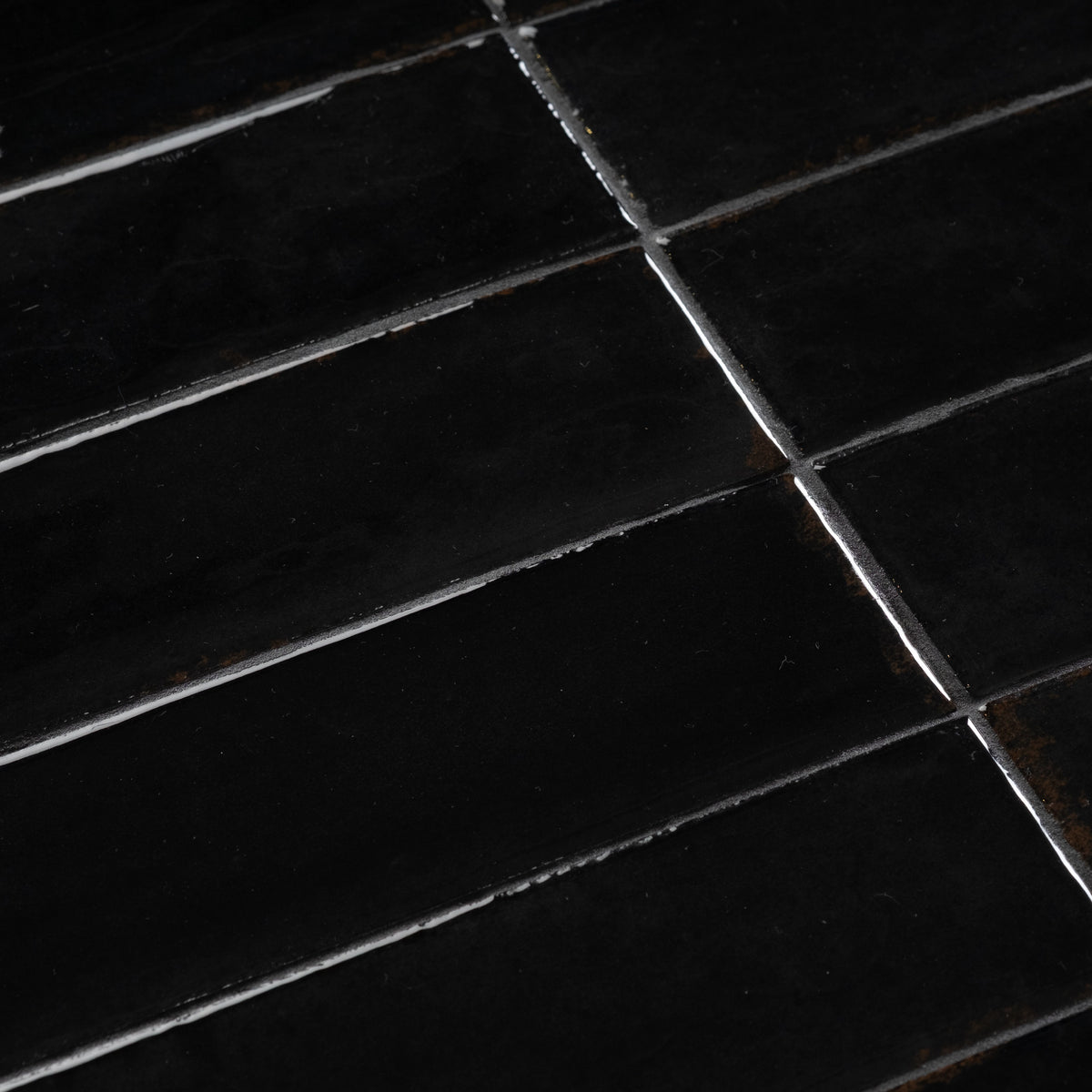 Close-up of the Bloor Coffee Table’s Black glossy tile top, showing bright highlights along the tile edges, offset grout joints, and faint rust-toned flecks.