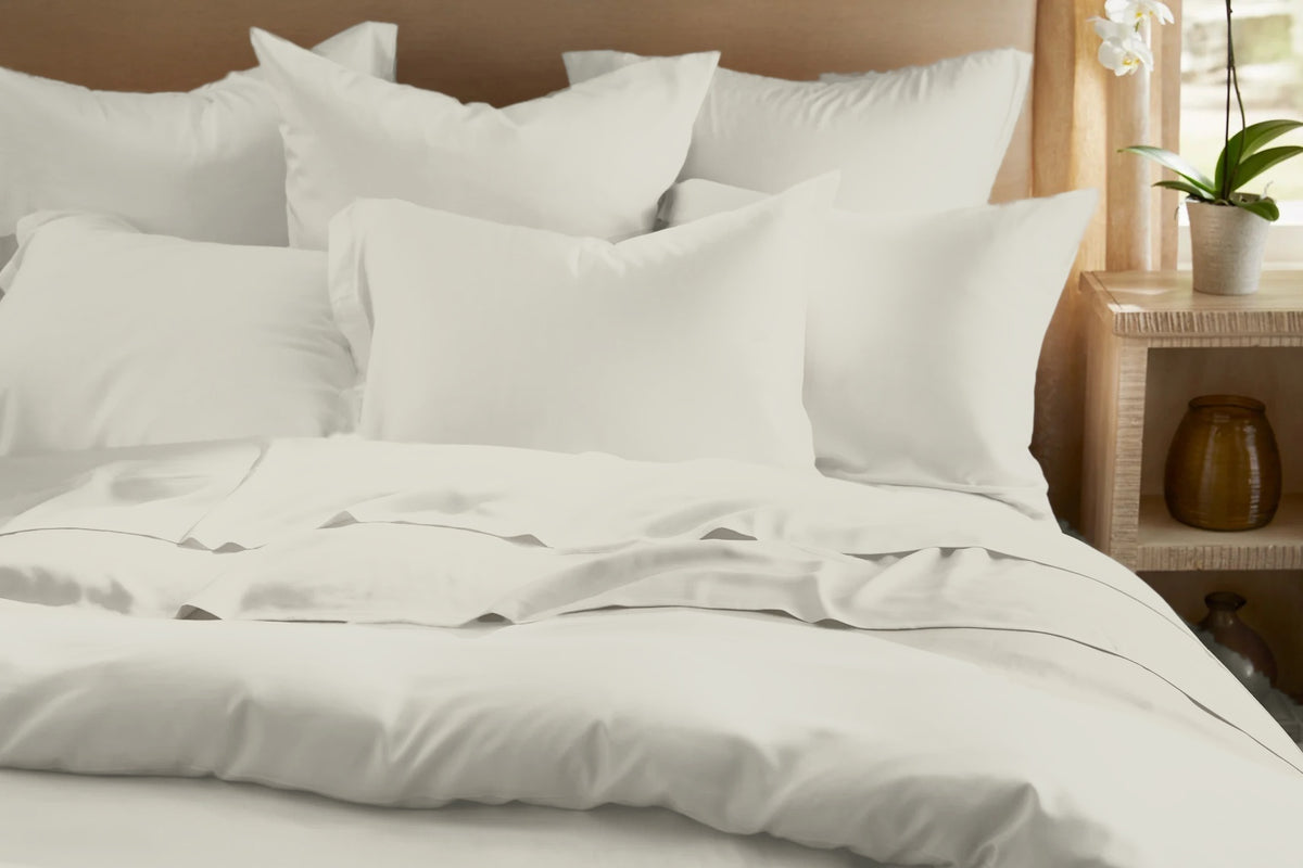 Legna Classic Bedding Set shown from the front in Sand, a soft ivory-beige with a gentle satin sheen, highlighting the layered pillow arrangement, flanged pillowcase edges, and the flat sheet folded over the duvet cove