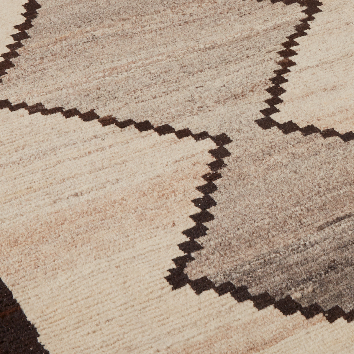 Close-up of a woven rug with a geometric pattern in beige, taupe, and dark brown, showing the texture and detail of the pile.