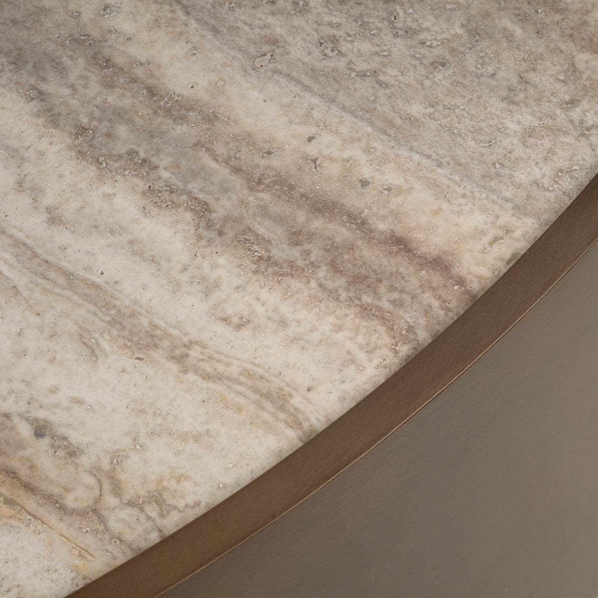 Close-up showing the edge of the Pedra Round Coffee Table’s cream travertine top with soft taupe banding and natural pitting, framed by a thin Aged Bronze metal rim.