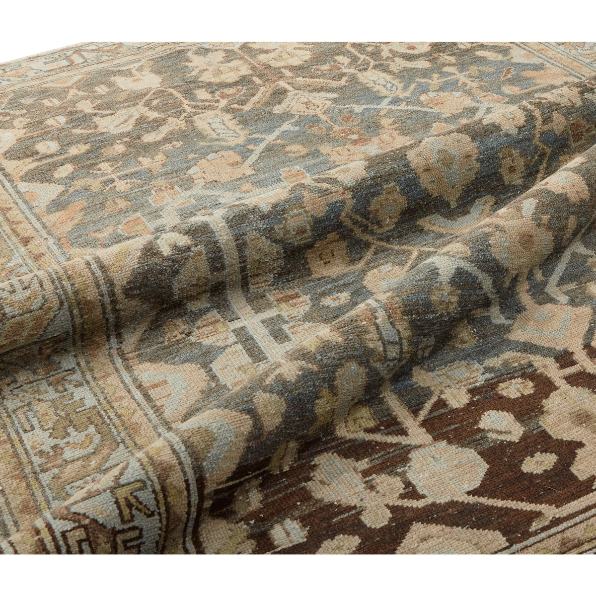 A close-up of a folded area rug showing its detailed traditional pattern in muted tones of beige, brown, slate blue, and cream.