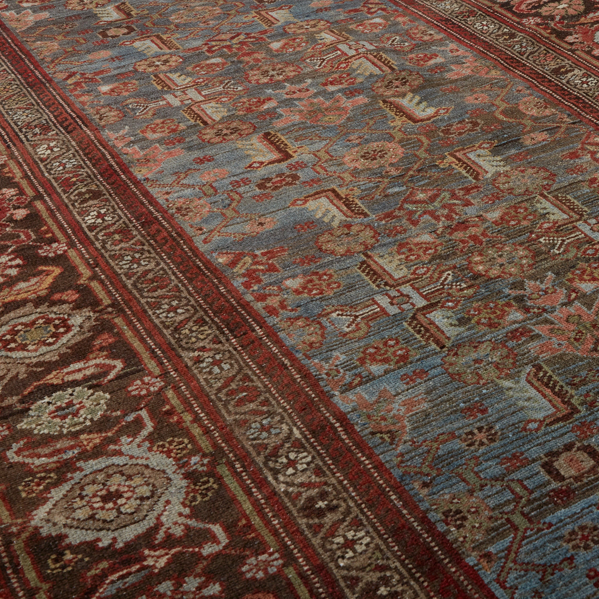 A close-up view of an ornate area rug with a traditional Persian-style pattern. The design features a dusty blue central field with red, beige, and brown floral motifs, bordered by deep red and dark brown sections with intricate geometric and floral details.