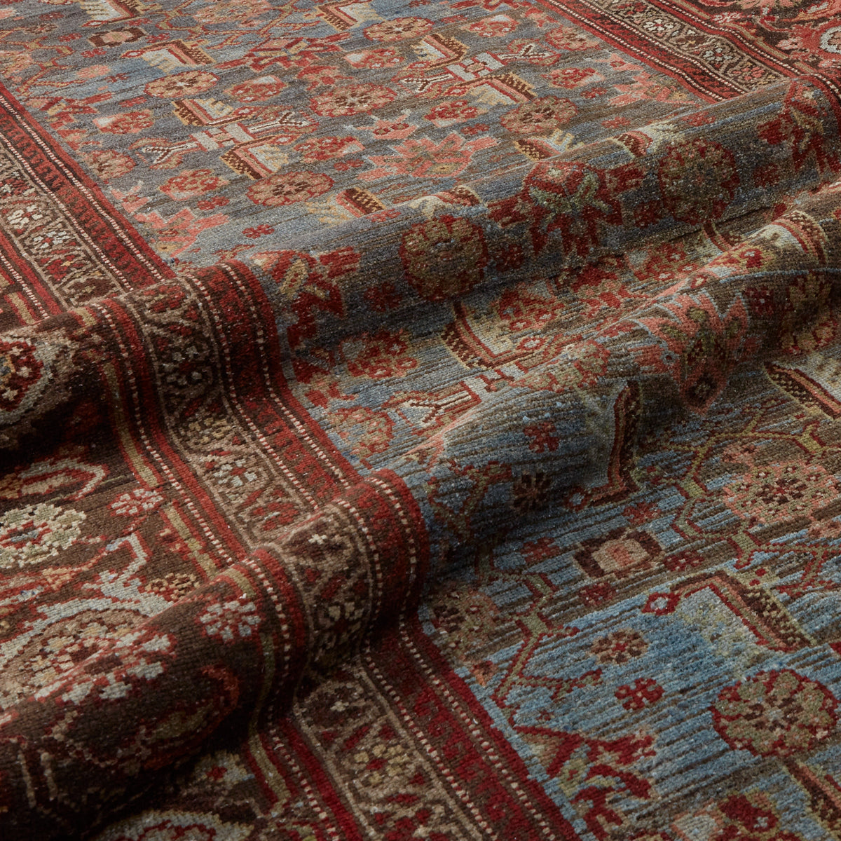 A close-up of a traditional patterned area rug with a dusty blue field, red borders, and brown motifs, shown with soft folds highlighting its texture.