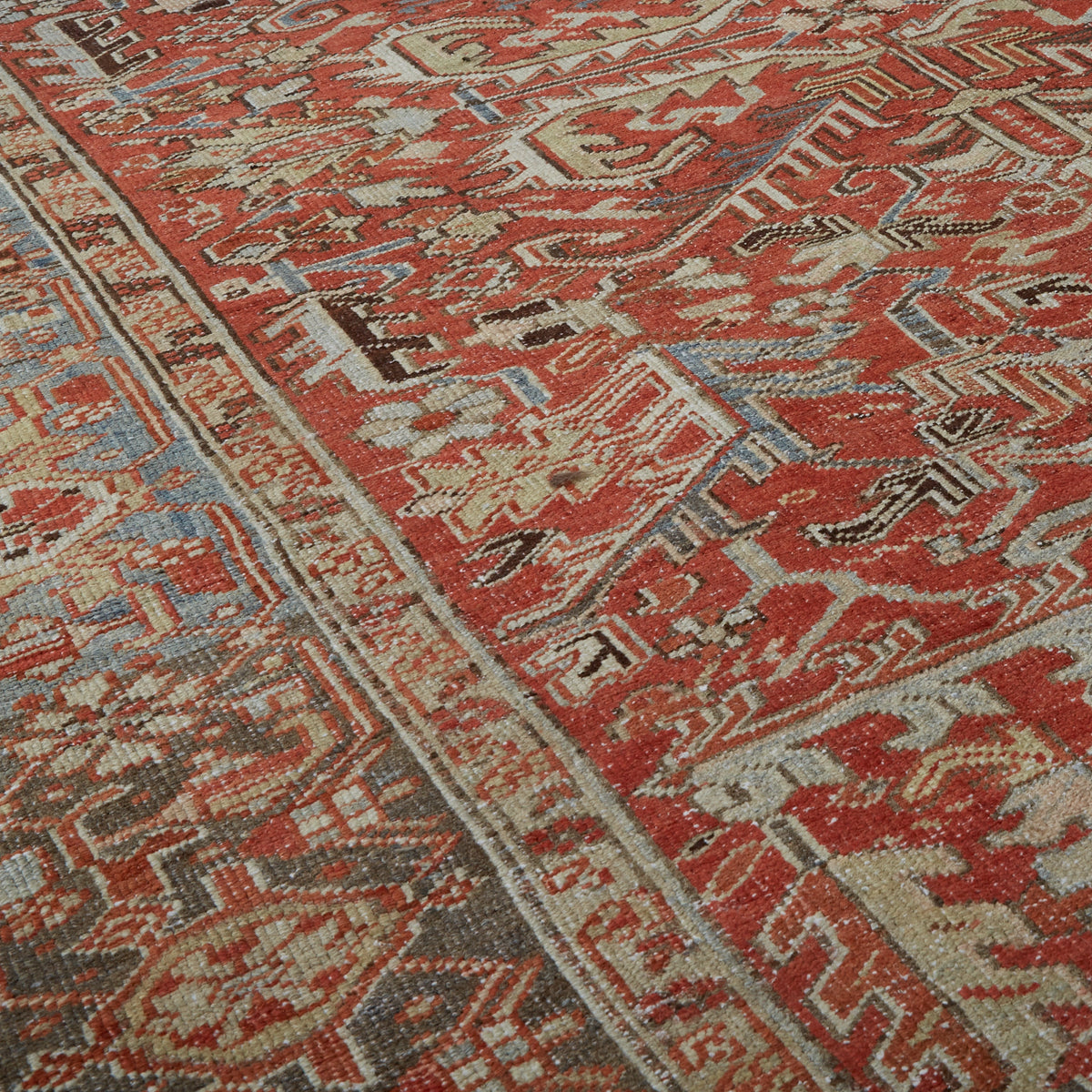 A close-up of a traditional patterned rug with a warm red-orange field, cream and beige geometric motifs, and blue-gray accents.