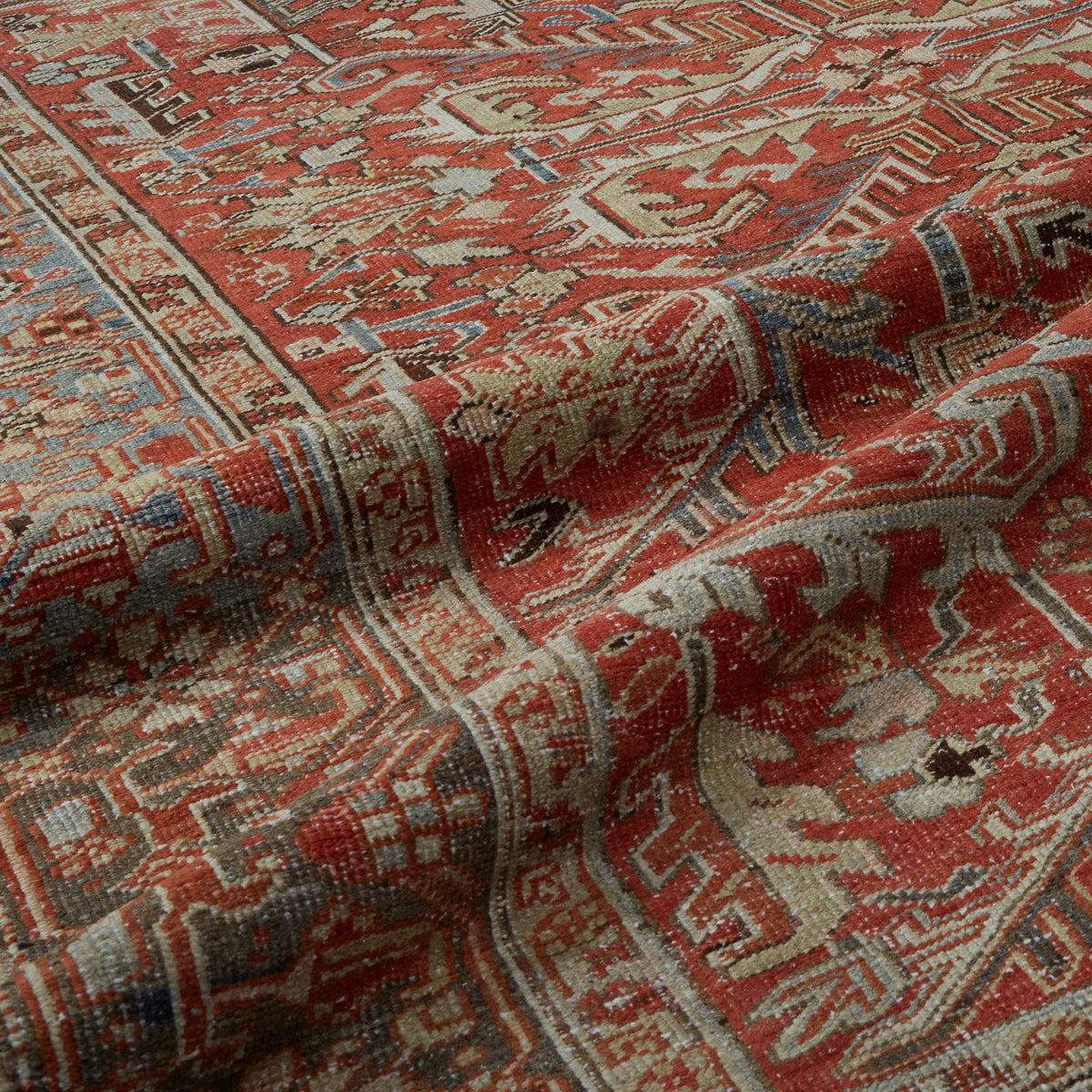 A close-up of a patterned area rug with a warm rust-red field, beige and cream geometric motifs, and accents of blue and gray.
